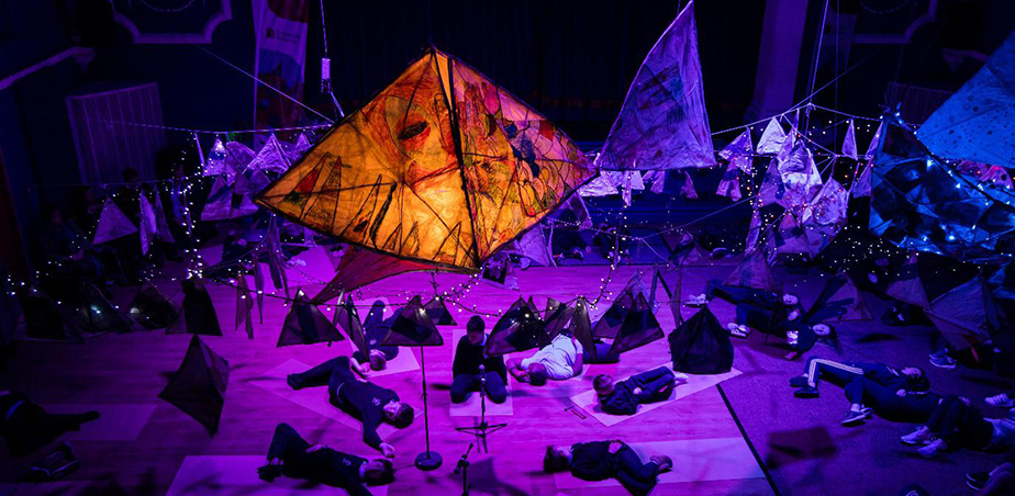 Orange paper fish sculpture hovering over purple stage with people lying down: Creative Schools Week 2019, Mullingar