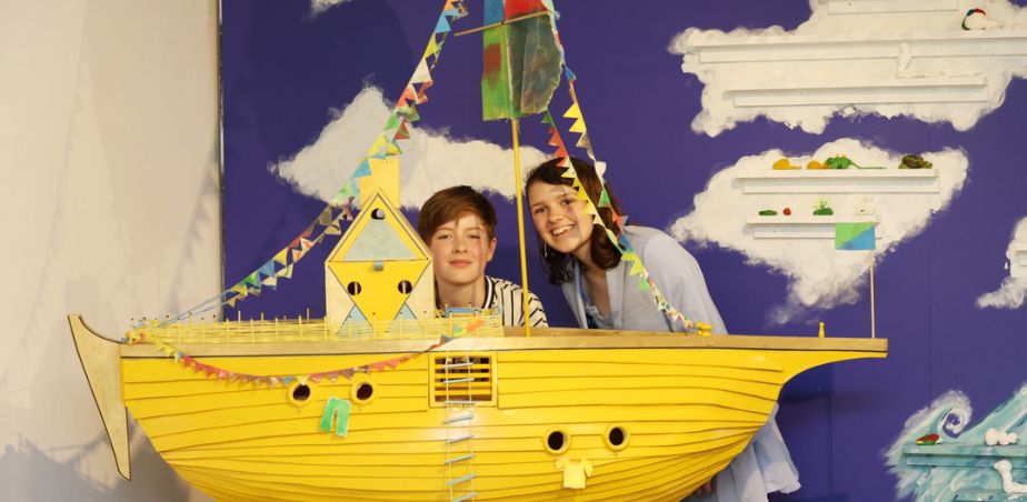 2 children smile from behind an artwork of a yellow boat